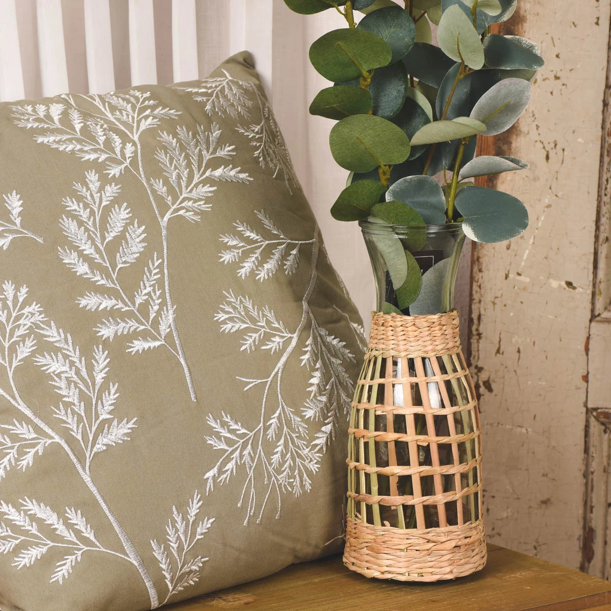 Grünes Kissen bestickt mit floralen Motiv neben einer Vase mit Rattangeflecht