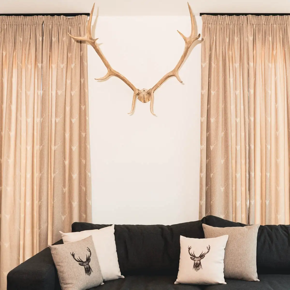 Wohnzimmer im Landhausstil: Beige Vorhänge mit Hirschmotiv, beige Kissen mit Hirschmotiv auf einem dunklen Sofa