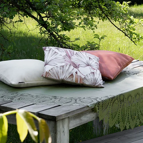 Drei elegante Outdoor-Kissen in Naturtönen liegen auf einem Holztisch mit Spitzen-Tischdecke unter einem Apfelbaum. Drei elegante Outdoor-Kissen in Naturtönen liegen auf einem Holztisch mit Spitzen-Tischdecke unter einem Apfelbaum.