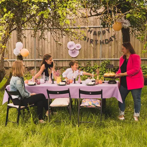 gartenparty mit gedecktem tisch mit genähter lilafarbener tischdecke aus der schöner leben manufaktur mit stabkerzen in kerzenhaltern, rosetten, einer happy birthday girlande und pompons in den bäumen