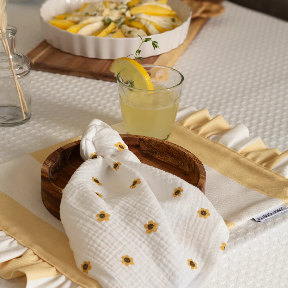 Sommerliche Tischdecke in Waffelstruktur mit Musselinserviette im Blümchenmuster, Holztablett und Erfrischungsgetränk – stilvolle Kombination für die warme Jahreszeit.