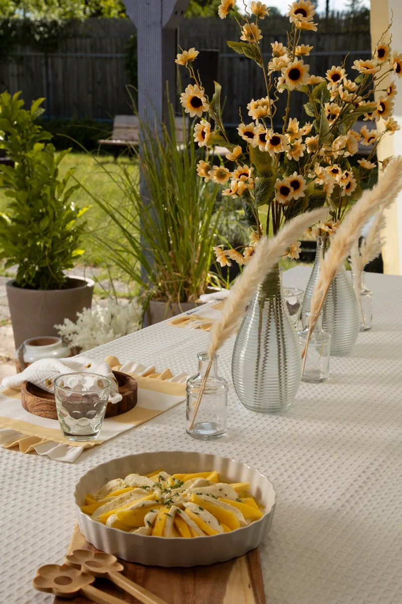 Detailaufnahme eines Gartentisches mit strukturierter Tischdecke, dekoriert mit Glasvasen, Sonnenblumen und Geschirr – natürliche Sommerdeko mit Stoffen in Gelbtönen.