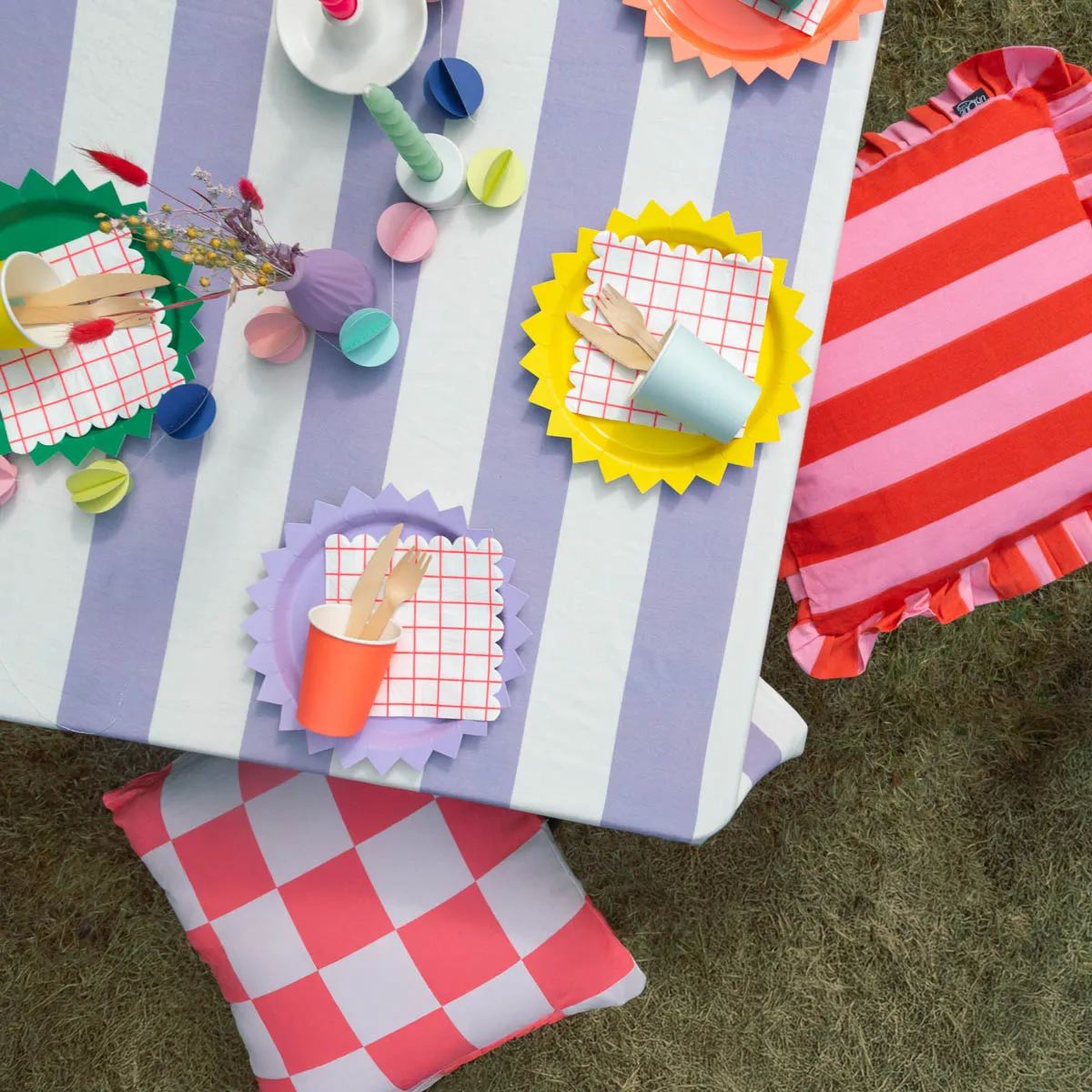 Gedeckter Tisch mit gestreifter Tischdecke in Lila-Weiß, bunten Tellern und Bechern, Papier-Servietten sowie Kissen in Rot-Pink und Rosa-Weiß – Sommerfest im Garten – SCHÖNER LEBEN