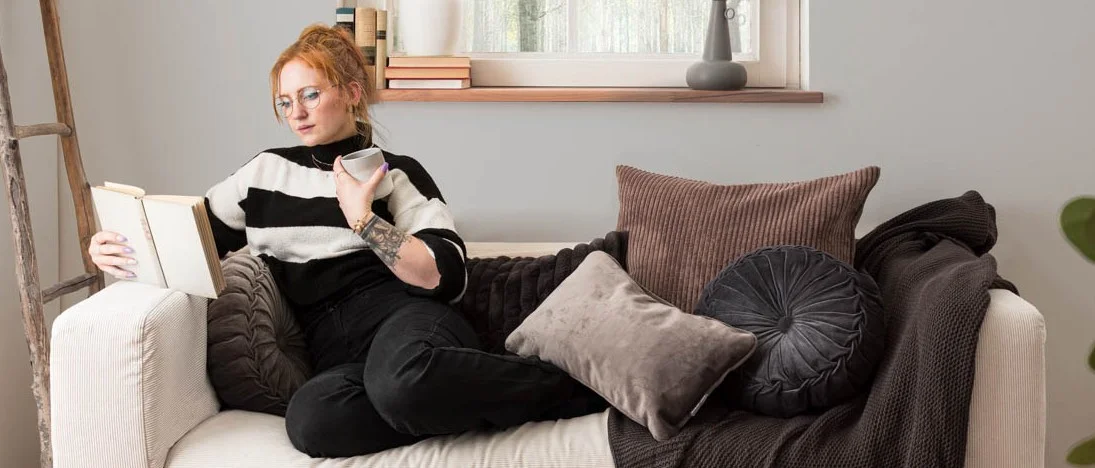 Frau sitz gemütlich auf einem Sofa mit Kissen, Decken. Im Hintergrund Gardinen und Wiohnaccessoires