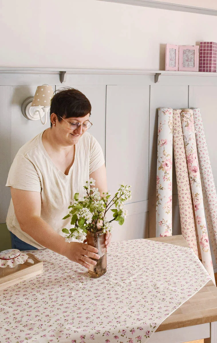 Frau stellt eine Vase mit frischen Blumen auf einen Tisch auf dem eine Tischdecke mit Rosenmotiv liegt
