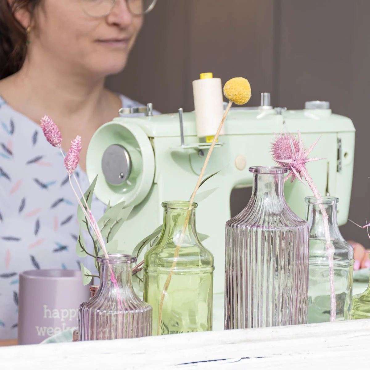 Glasvasen in verschiedenen Größen und in verschiedenen Farben stehen vor einer Pastellgrünen Nähmaschine Glasvasen in verschiedenen Größen und in verschiedenen Farben stehen vor einer Pastellgrünen Nähmaschine