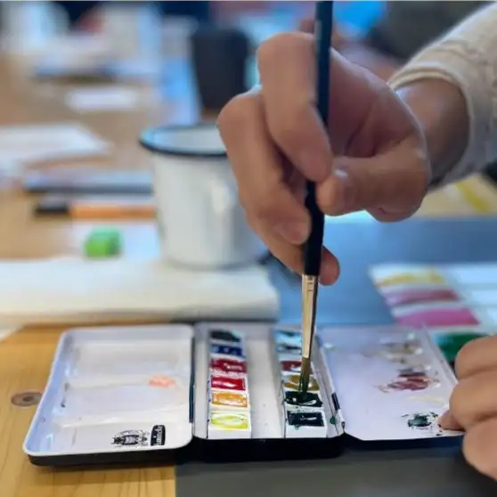 Hand mit Pinsel, die Farbe aus einem Aquarellmalkasten aufnimmt Hand mit Pinsel, die Farbe aus einem Aquarellmalkasten aufnimmt