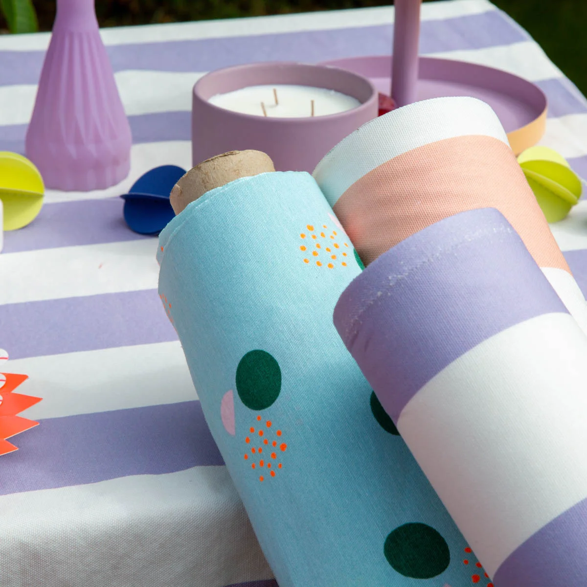 Dreiei bunte Stoffrollen in sommerlichen Farben auf einem gestreiften Tisch mit Kerzen und Dekoration – ideal für kreative DIY-Projekte mit Baumwollstoffen