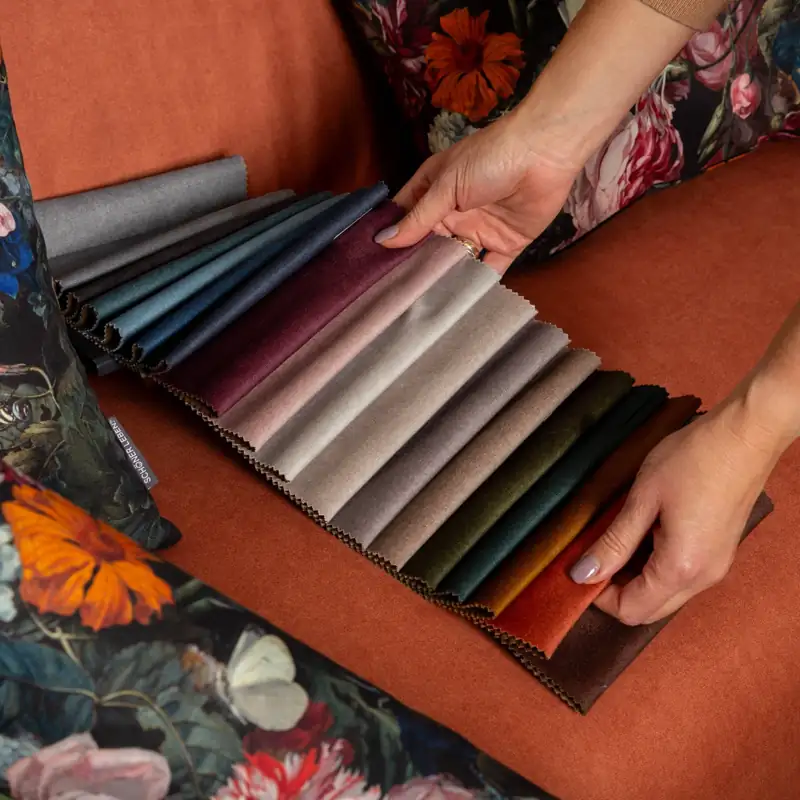 Fächer mit Polsterstoffen in verschiedenen Farben auf einem Sessel mit floralen Kissen – hochwertige Möbelstoffe als Meterware zum Polstern und Beziehen