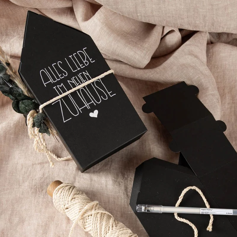 Schwarze Geschenkverpackung in Hausform mit der weißen Aufschrift „ALLES LIEBE IM NEUEN ZUHAUSE“ und einem kleinen Herz darunter. Die Verpackung liegt auf einem beigen Leinenstoff, daneben Bastelmaterialien wie Juteschnur, ein weißer Gelstift und eine weitere ungefaltete schwarze Kartonverpackung.