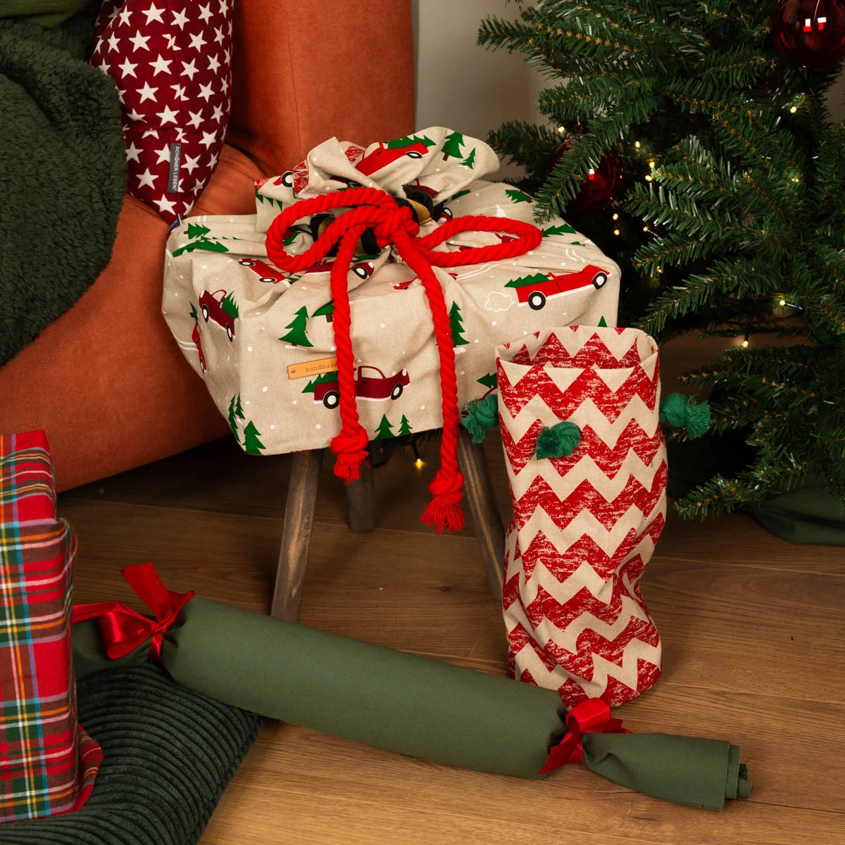 Kreativ verpackte Geschenke mit Weihnachtsstoffen: ein beiger Stoff mit roten Trucks und Tannenbäumen, verziert mit einer roten Kordel, sowie ein roter Chevron-Musterstoff. Im Vord Kreativ verpackte Geschenke mit Weihnachtsstoffen: ein beiger Stoff mit roten Trucks und Tannenbäumen, verziert mit einer roten Kordel, sowie ein roter Chevron-Musterstoff. Im Vord