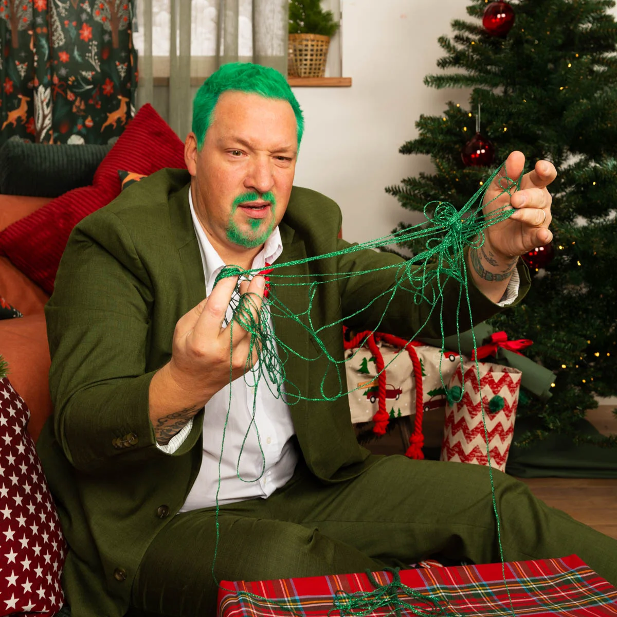 Ein Mann mit grün gefärbtem Haar und Bart in einem grünen Anzug kämpft mit verworrenen grünen Schnüren, während er Geschenke mit Weihnachtsstoff einpackt. Im Hintergrund sind ein g Ein Mann mit grün gefärbtem Haar und Bart in einem grünen Anzug kämpft mit verworrenen grünen Schnüren, während er Geschenke mit Weihnachtsstoff einpackt. Im Hintergrund sind ein g
