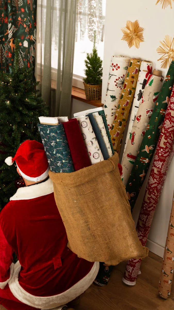 Eine Person im Weihnachtsmannkostüm trägt einen großen Jutesack voller Stoffrollen mit weihnachtlichen Designs: blaue Stoffe mit Schneeflocken, dunkelrote Meterware und weiße Stoff Eine Person im Weihnachtsmannkostüm trägt einen großen Jutesack voller Stoffrollen mit weihnachtlichen Designs: blaue Stoffe mit Schneeflocken, dunkelrote Meterware und weiße Stoff