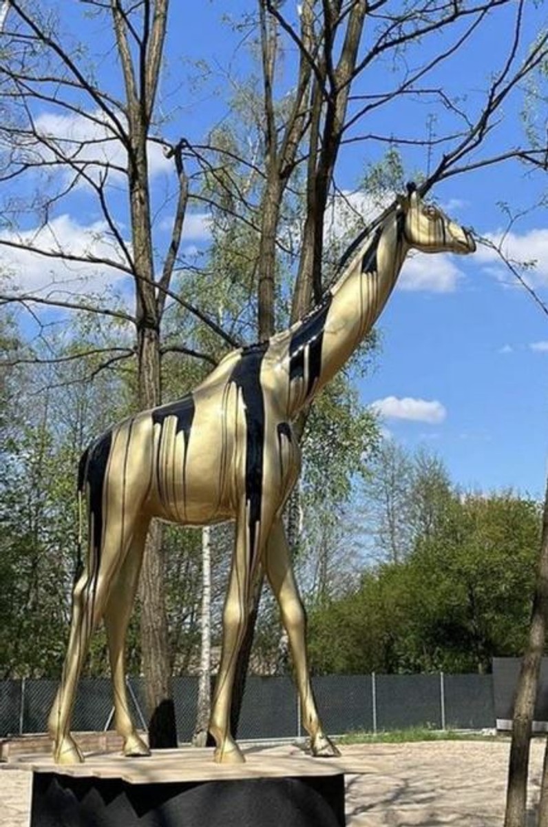 Vous voyez une sculpture de girafe XXL grandeur nature dans les couleurs or et noir. La sculpture est dans un parc.