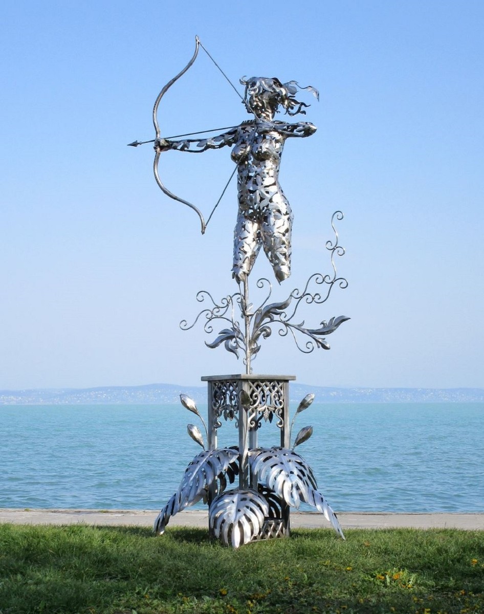 Vous voyez une figurine en acier inoxydable faite à la main en forme d’archère tirant un arc. La base sur laquelle repose la figurine est décorée dans un style floral ondulé. En arrière-plan, on aperçoit un plan d’eau.