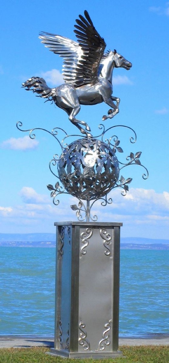 Vous voyez une figurine en acier inoxydable faite à la main en forme de Pégase qui se tient sur un ballon et vole. La base sur laquelle repose la figurine est dans un style ondulé et élégant en acier inoxydable. En arrière-plan, on aperçoit un plan d’eau.