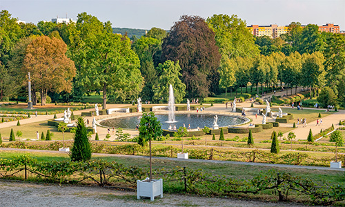 Capture d'image panoramique du parc municipal de Potsdam avec une fontaine centrale entourée de fontaines d'eau bouillonnante, de sculptures ornées, de statues et de plantes ornementales.