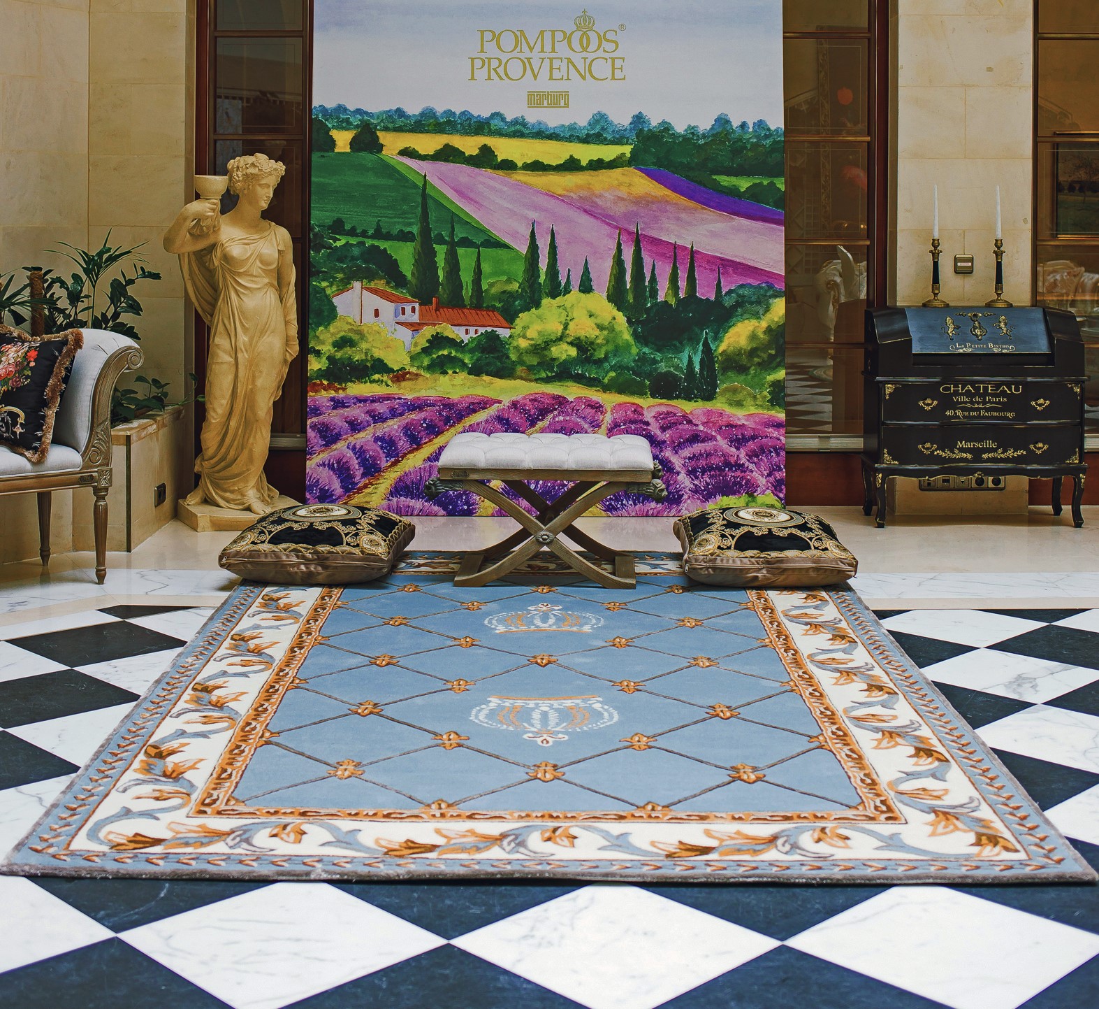 En la imagen se puede ver la nueva colección de Harald Glööckler Pompöös Provence, el suelo con diseño de tablero de ajedrez en blanco y negro, la alfombra azul cielo con corona, los cojines de los asientos y la mesa al fondo, un cartel publicitario que muestra la Provenza del sur de Francia.