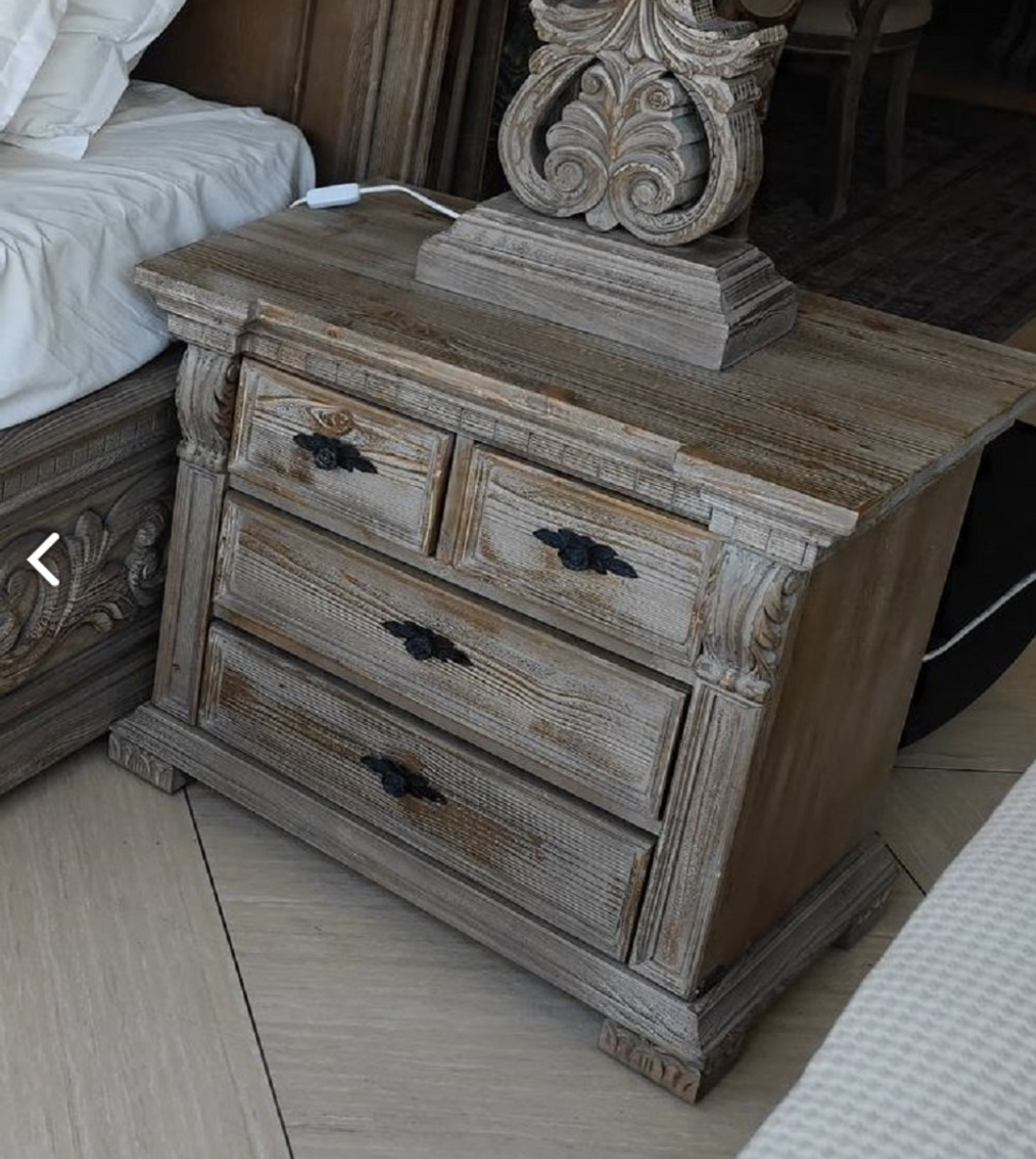 You see a luxurious hand-carved bedside table with two large and two small drawers from the Harald Glööckler Pompöös Provence collection 2024 in a baroque country house style in the colors brown gray made of solid wood and metal handles.