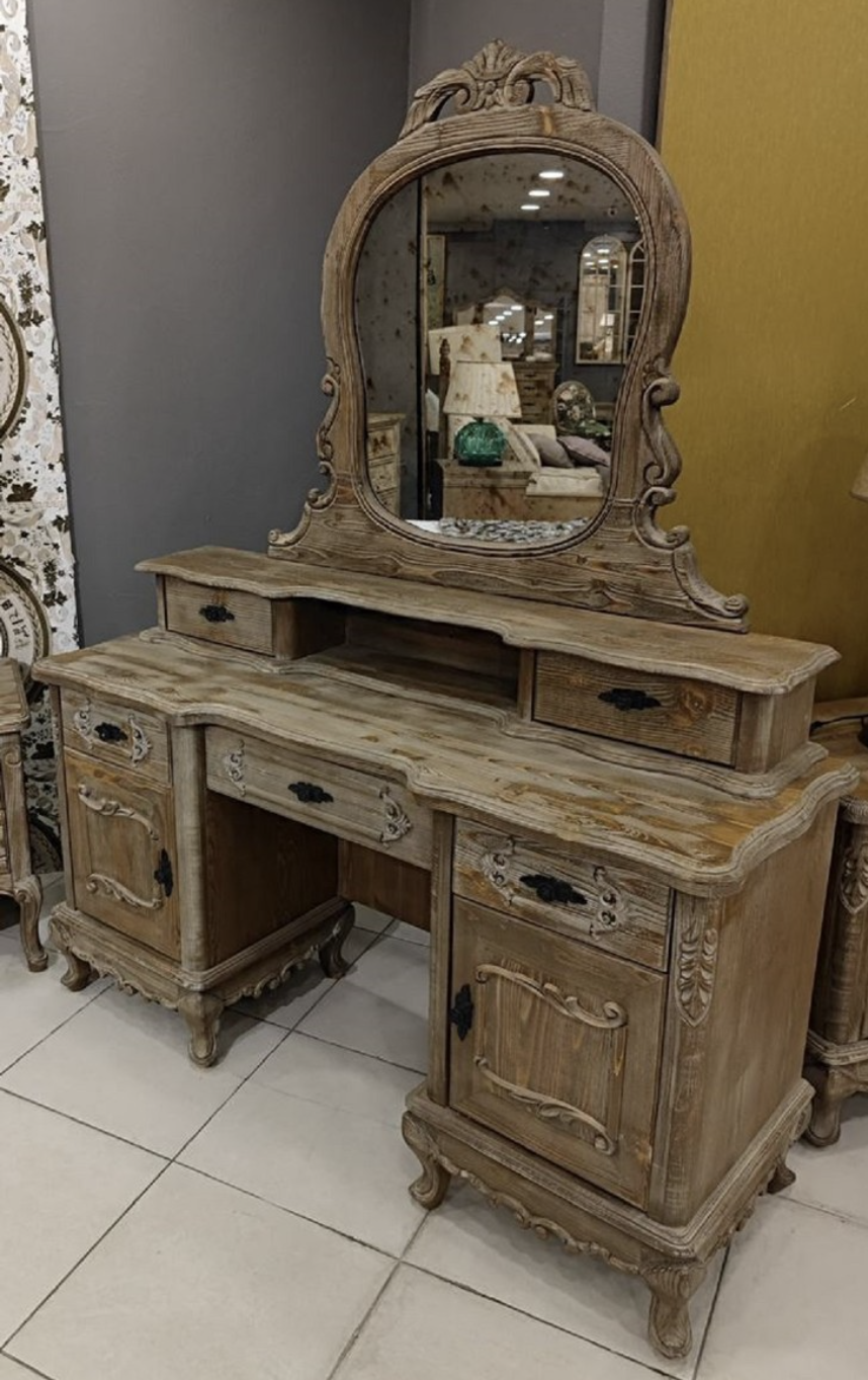 Vous voyez une luxueuse coiffeuse sculptée à la main avec miroir de la collection Harald Glööckler Pompöös Provence 2024 dans un style maison de campagne baroque dans les couleurs marron gris en bois massif.