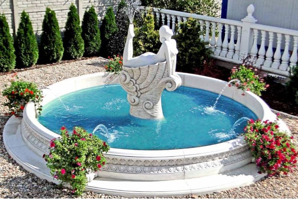 Vous voyez une fontaine très élégante avec quatre sorties d'eau dans l'entourage et une sculpture féminine allongée aux pieds de laquelle s'élève une fontaine d'eau. Fontaine couleur blanc.